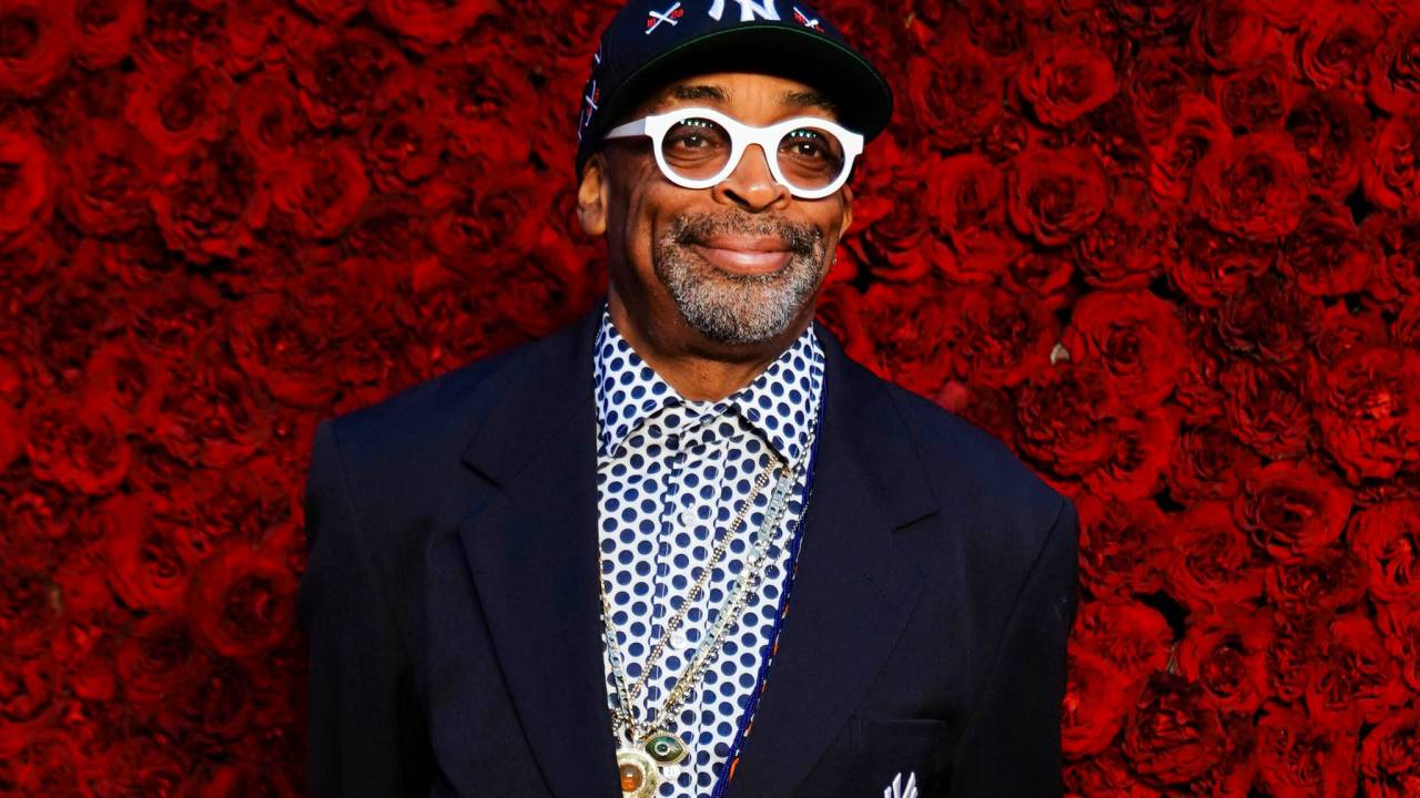 Foto mostra o diretor Spike Lee usando óculos escuros de aro branco, boné, camisa quadriculada azul e paletó escuro. O fundo da imagem tem rosas vermelhas