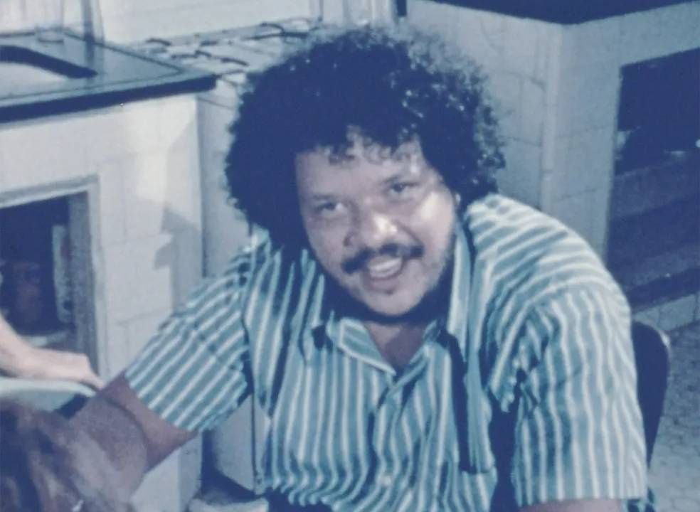 Tim Maia sorridente, de camisa listrada, com cabelo black power e bigode, em fotografia sépia.