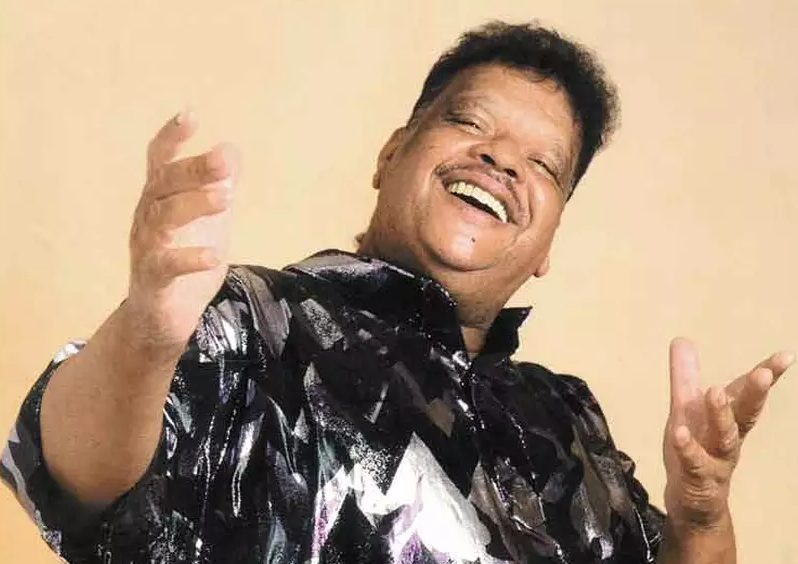 Tim Maia por volta de seus 50 anos, de blusa prateada e preta, sorrindo, com os braços apontando para a frente.