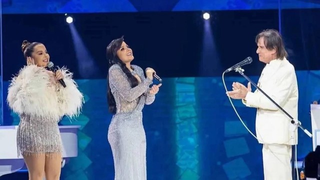 Maiara está de saia prateado de paetês e casaco branco de plumas, Maraisa está de vestido prateado de paetês e Roberto está de terno branco, cantando.