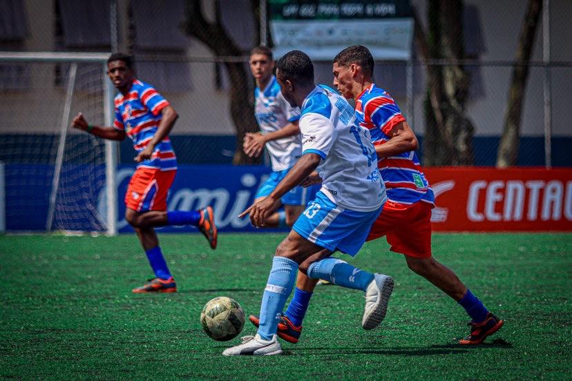 Foto mostra meninos disputando bola em jogo de futebol. Um usa uniforme azul claro e branco e outro azul mais escuro e vermelho