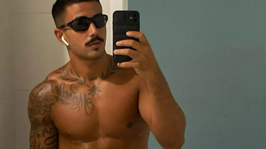 Yuri Meirelles é branco, usa bigode, tem cabelo curto castanho, está sem camisa, com fone sem fio, de óculos escuros e segurando o celular em uma foto do espelho.