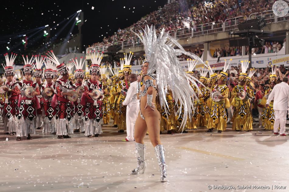 Paolla Oliveira usa o corpo coberto por detalhes prateados, além de ostentar um arranjo de cabeça prateado e botas na mesma cor. Ao fundo, a bateria da Grande Rio.