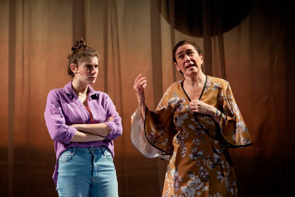 GIULIA BERTOLLI e LILIA CABRAL na peça A LISTA Credito Priscila Prade (10) Giulia está à esquerda, ela é uma mulher branca e está vestida com calça jeans, camisa lilás de manga e cabelo preso em coque. Ela tem os braços cruzados. À direita, Lilia, também branca, que usa um vestido bege estampado que lembra um quimono japonês. Ela está gesticulando.
