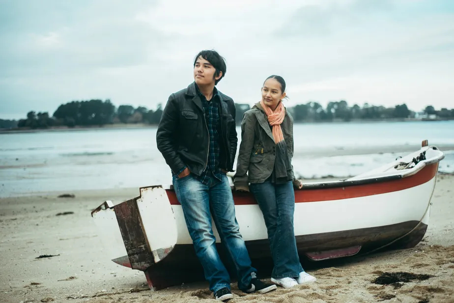 Os irmãos Isaac e Nora encostados em um pequeno barco na areia. Ao fundo, um rio.
