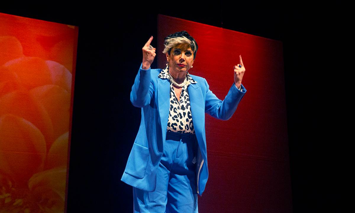 Heloisa Périssé está em cima de um palco com fundo vermelho, de terno azul e com camisa de oncinha discursando.