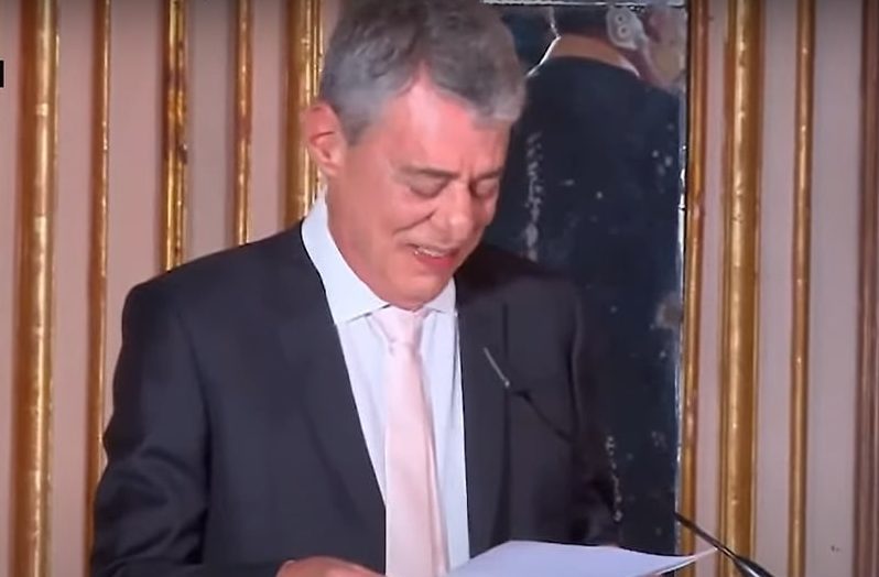Chico Buarque é branco, tem cabelo grisalho. Ele usa terno escuro com gravata amarelo-clara e está lindo um discurso com um leve sorriso no rosto.