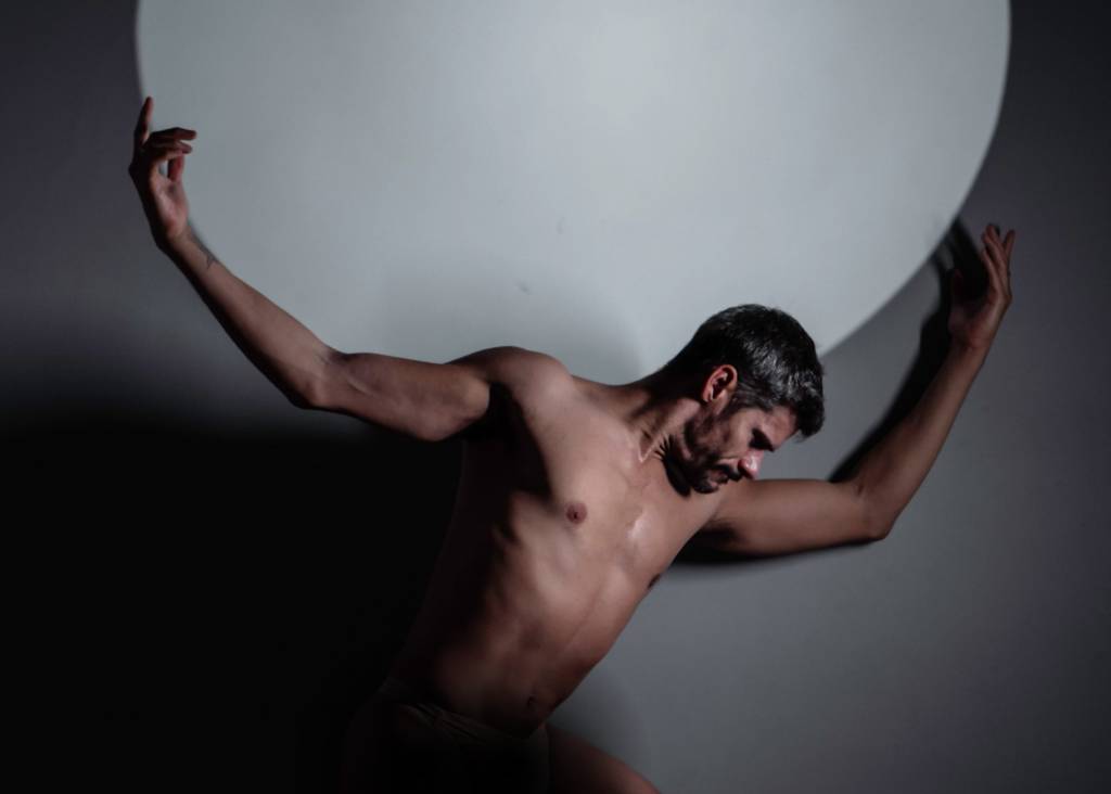 Thiago Soares está sem camisa e segura uma esfera branca nas costas, em foto com luz e sombra