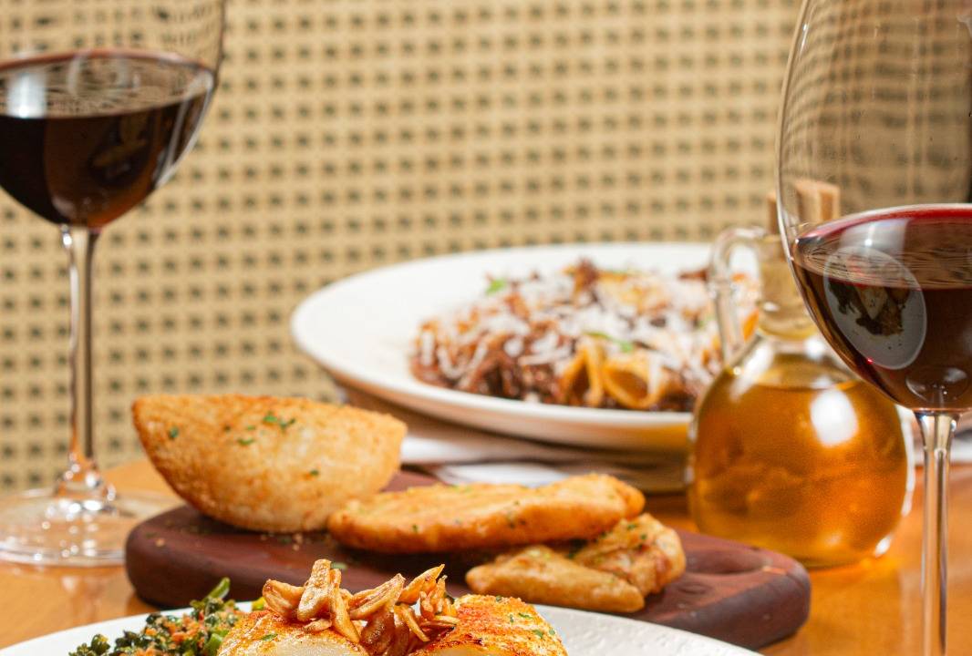 Foto mostra mesa com prato de bacalhau e duas taças de vinho tinto