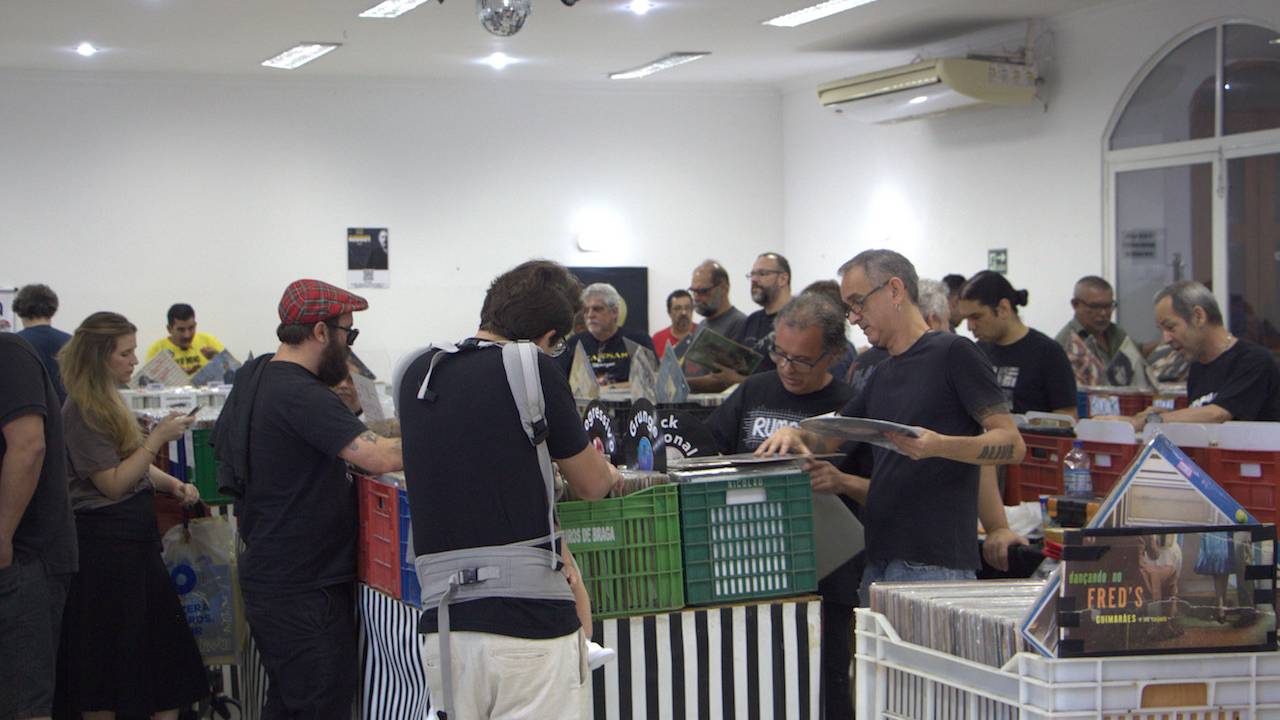Sala com engradados cheios de discos de vinil e pessoas procurando álbuns nelas
