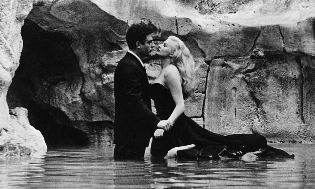 Foto em preto e branco com homem branco de cabelos escuros e terno e mulher loura platinada de vestido tomara que caia escuro prestes a se beijar, com as pernas submersas em uma fonte, com a parede de pedra ao fundo