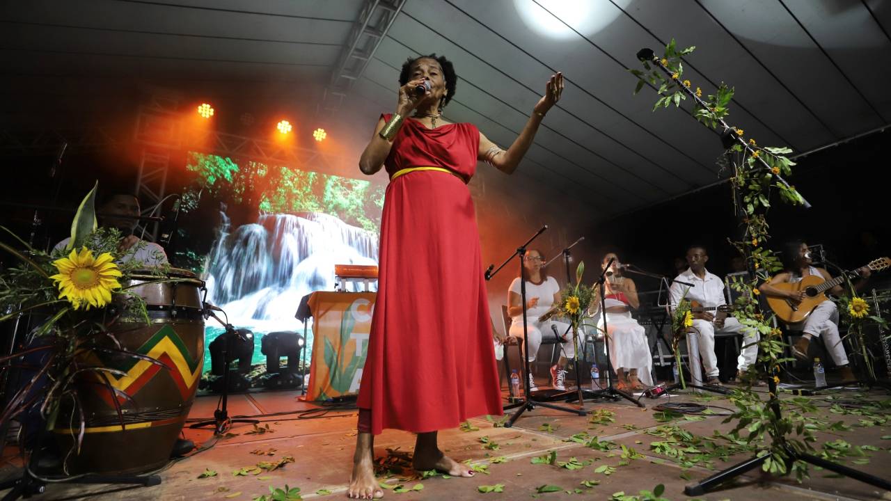 Laura Mariá é uma mulher negra, está de vestido vermelho, descalça, pisando em folhas, cantando. Ao fundo da foto, músicos sentados, vestidos de branco