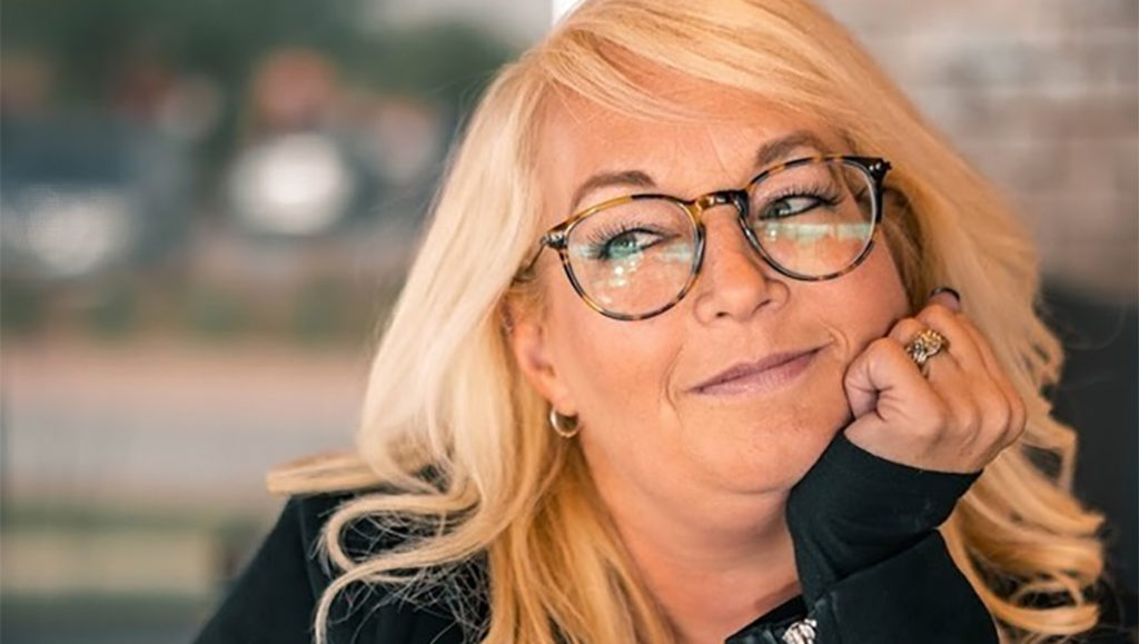 Lynn Painter é uma mulher branca, de cabelo comprido louro e liso, está de óculos de grau pretos e usa parte de cima preta