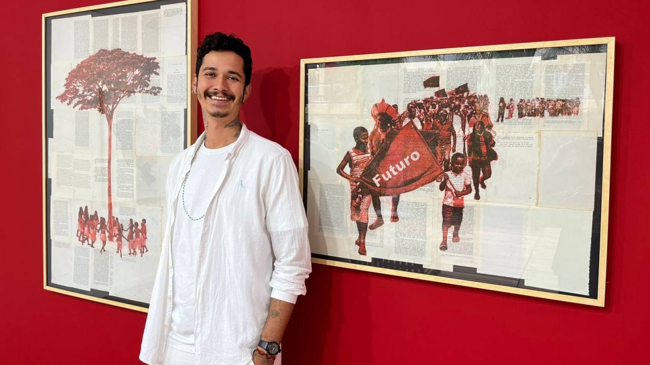 Hal Widson é um homem não branco de pele clara, cabelos e bigode pretos. Ele usa camisa, blusa e calça brancas, tem as mãos nos bolsos e sorri. Ao fundo, duas obras suas, com pinturas sobre páginas de livros coladas: um quadro uma árvore e cercada por pessoas, em fotografia em preto e branco recortada e pintada de vermelho, e a outra uma marcha de pessoas, feita com fotografia em preto e branco recortada e pintada de vermelho, em que elas carregam uma bandeira onde se lê futuro.