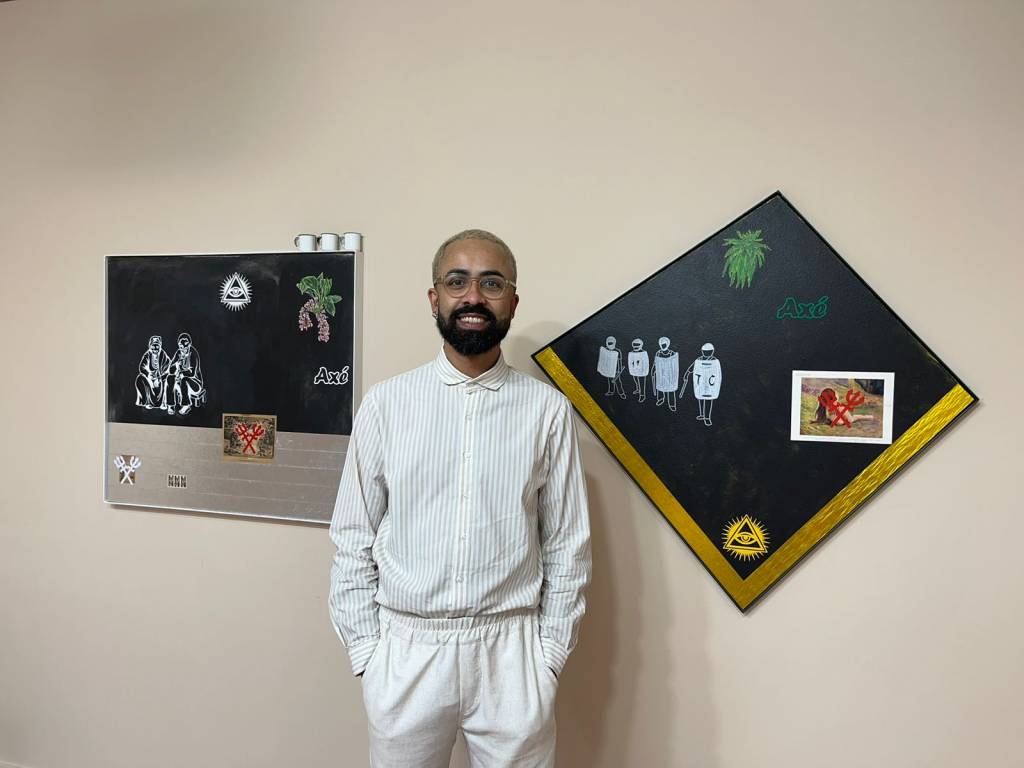 jaime lauriano Jaime Lauriano é um homem negro de pele clara. Ele está de camisa branca listrada e calça branca, sorrindo e com as mãos no bolso. Tem o cabelo curto e descolorido, barba, bigode e cavanhaque pretos. Atrás dele, estão dois quadros: um é retangular e o outro é quadrado, com um dos vértices para cima. Os dois com fundo preto.