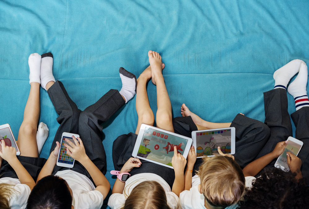 Crianças sobre uma cama brincando com tablets e celulares.