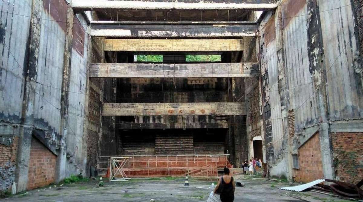 Interior de um pr&eacute;dio abandonado com paredes de concreto e tijolos expostos, vigas de madeira e metal enferrujadas. Uma mulher de costas, com blusa escura, caminha em primeiro plano. Ao fundo, um grupo de pessoas e uma cerca laranja. O local parece deteriorado, com vegeta&ccedil;&atilde;o rasteira e entulho no ch&atilde;o