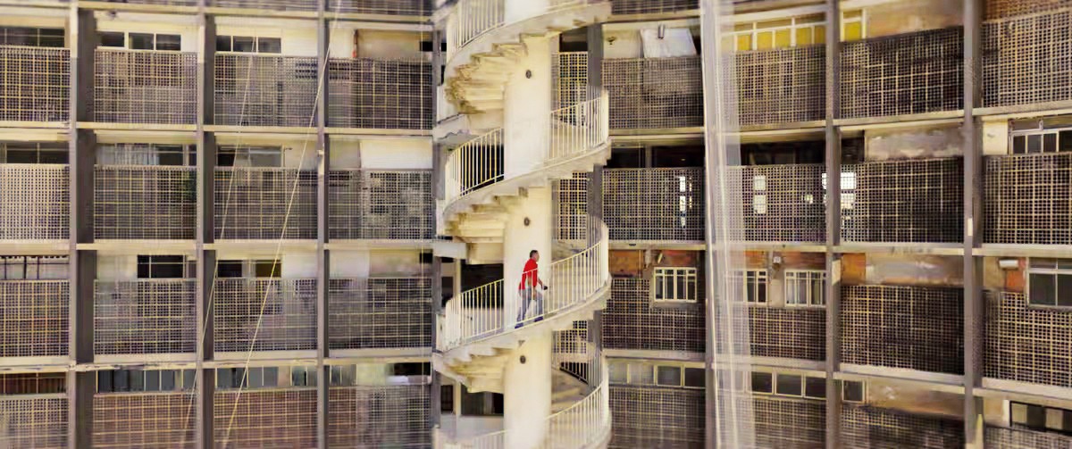 Homem de camiseta vermelha e cal&ccedil;a cinza descendo uma escada caracol branca em um pr&eacute;dio de apartamentos com janelas gradeadas