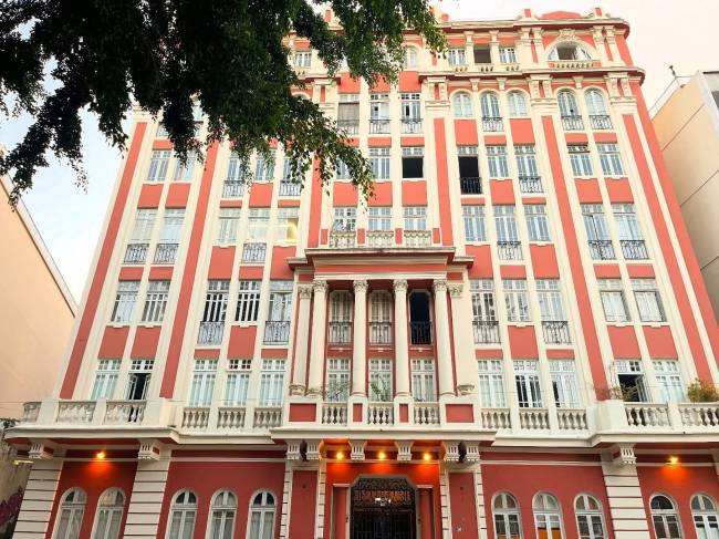 Edif&iacute;cio alto com fachada em tons de vermelho e creme, janelas retangulares e sacadas com grades, sob um c&eacute;u claro e galhos de &aacute;rvore no canto superior esquerdo