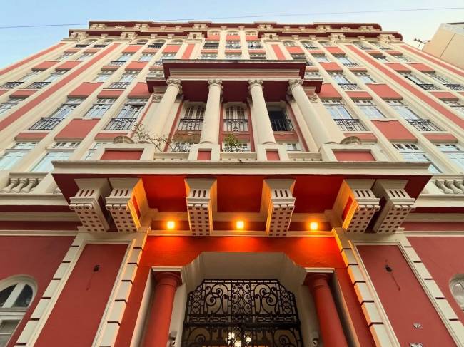 Fachada de um edif&iacute;cio alto com detalhes arquitet&ocirc;nicos em vermelho e creme, visto de baixo para cima. A entrada tem um port&atilde;o de ferro trabalhado e colunas vermelhas, com luzes acesas no teto da marquise. As janelas s&atilde;o retangulares, com sacadas em ferro forjado. O c&eacute;u azul claro &eacute; vis&iacute;vel ao fundo