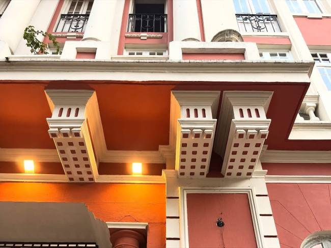 Detalhe da fachada de um pr&eacute;dio com paredes brancas e detalhes em terracota. Abaixo de uma sacada, h&aacute; um teto laranja com tr&ecirc;s suportes brancos ornamentados e duas luzes quadradas acesas
