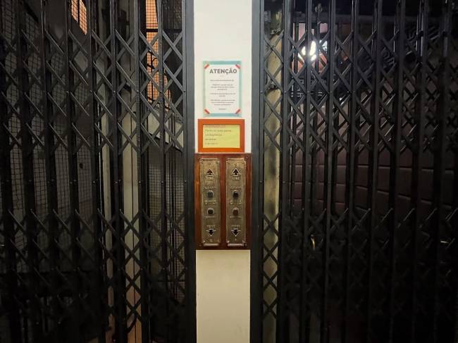 Porta de elevador antiga com grades met&aacute;licas sanfonadas pretas, painel de madeira e bot&otilde;es de lat&atilde;o. Acima, avisos de 