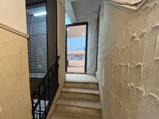 Escadaria interna de um pr&eacute;dio antigo com paredes texturizadas em tom bege, corrim&atilde;o de ferro preto e uma porta aberta ao fundo revelando um ambiente externo iluminado