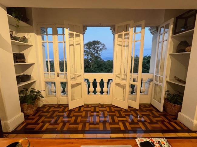 Varanda com portas francesas brancas abertas para uma vista de &aacute;rvores e c&eacute;u azul claro, piso de madeira em padr&atilde;o geom&eacute;trico e estantes brancas nas laterais com objetos decorativos e plantas