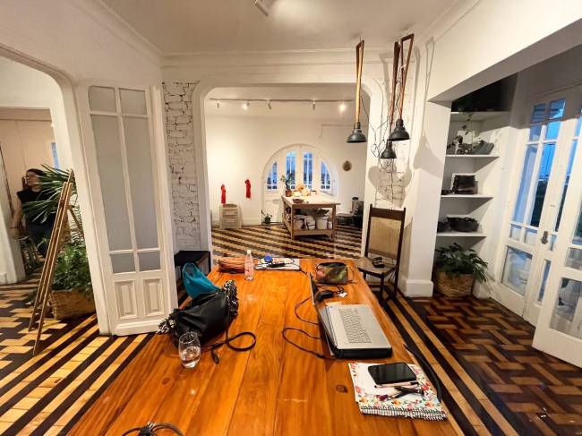 Mesa de madeira r&uacute;stica com notebook aberto, celular, garrafa de &aacute;gua e bolsa, em ambiente interno com piso de madeira em zigue-zague e listras, paredes brancas, arcos e portas brancas. Ao fundo, uma pessoa e uma ilha de cozinha.