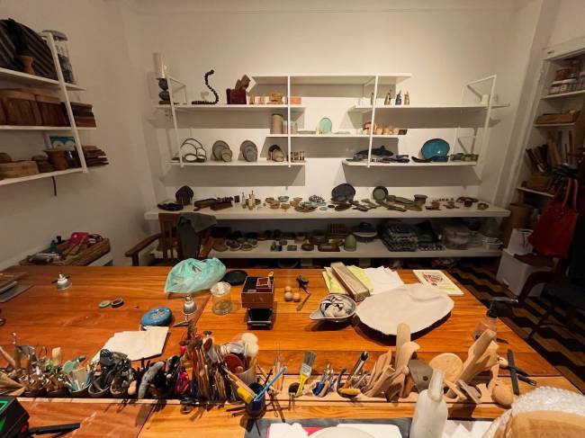 Ateli&ecirc; de artesanato com uma mesa de madeira cheia de ferramentas e materiais, como pinc&eacute;is, tintas, pe&ccedil;as de cer&acirc;mica e madeira, e prateleiras brancas ao fundo exibindo diversos objetos artesanais