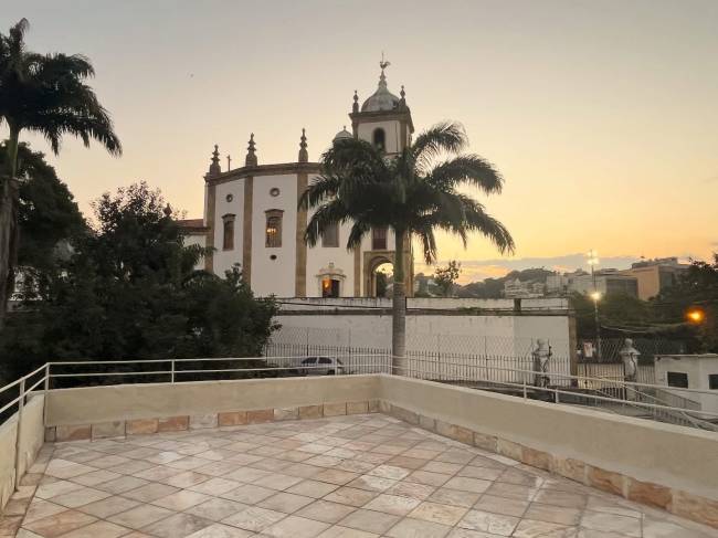 Terra&ccedil;o com piso de cer&acirc;mica e guarda-corpo branco, tendo ao fundo uma igreja colonial branca com torre sineira e palmeiras, sob um c&eacute;u alaranjado de p&ocirc;r do sol