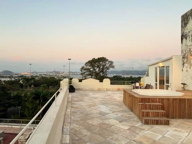 Terra&ccedil;o com piso de pedra, banheira de hidromassagem em deck de madeira e vista para uma cidade costeira com montanhas ao fundo, sob c&eacute;u claro ao entardecer