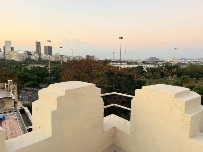 Vista panor&acirc;mica do Rio de Janeiro ao entardecer, com pr&eacute;dios altos no horizonte, &aacute;rvores verdes e um c&eacute;u em tons de rosa e azul, emoldurada por uma mureta de concreto em primeiro plano