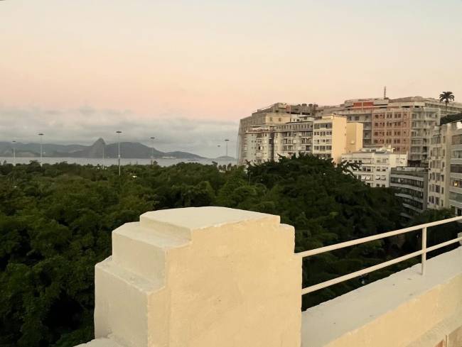 Vista panor&acirc;mica do Rio de Janeiro ao entardecer, com o P&atilde;o de A&ccedil;&uacute;car ao fundo, uma vasta &aacute;rea verde, pr&eacute;dios residenciais e um parapeito branco em primeiro plano