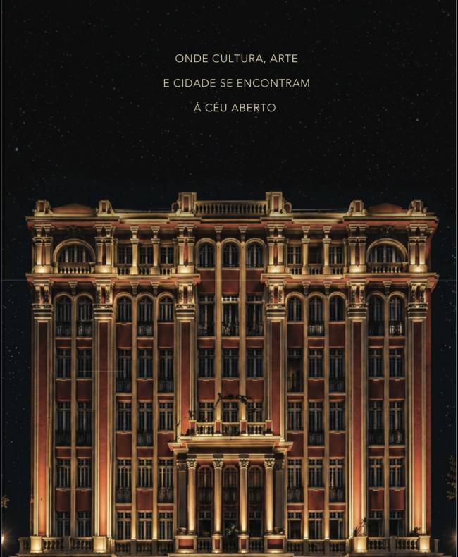 Edif&iacute;cio hist&oacute;rico imponente de cor terracota com detalhes arquitet&ocirc;nicos cl&aacute;ssicos e colunas iluminadas por luzes quentes, sob um c&eacute;u noturno estrelado. O texto branco no topo diz: 