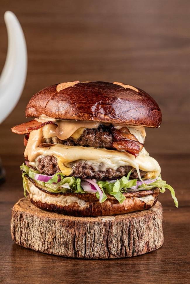 Hamb&uacute;rguer duplo com p&atilde;o brioche escuro, dois hamb&uacute;rgueres de carne, queijo derretido, bacon crocante, alface e cebola roxa, servido sobre um tronco de madeira r&uacute;stico em uma mesa escura