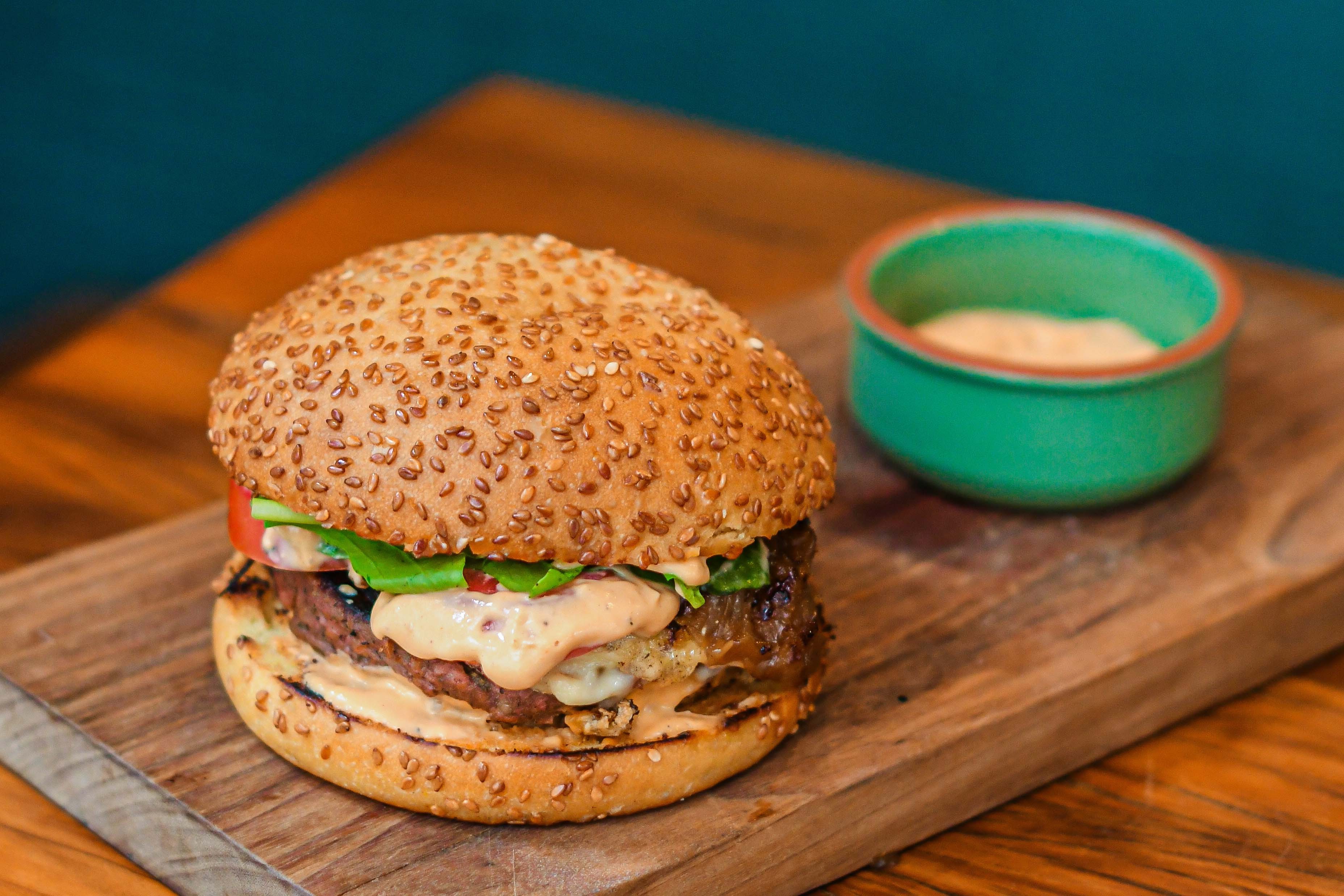 Hamb&uacute;rguer com p&atilde;o de gergelim, alface, tomate, carne e molho cremoso, servido em t&aacute;bua de madeira com um pote verde de molho ao lado