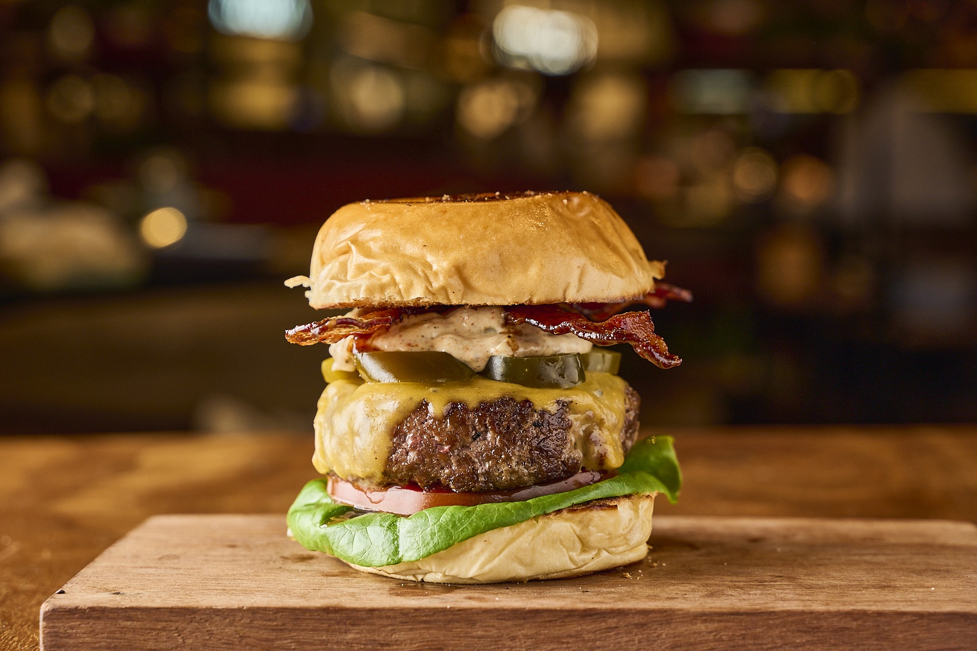 Hamb&uacute;rguer gourmet com p&atilde;o brioche, bacon crocante, molho cremoso, picles, queijo derretido, carne, tomate e alface, servido sobre uma t&aacute;bua de madeira em ambiente de restaurante com luzes desfocadas ao fundo