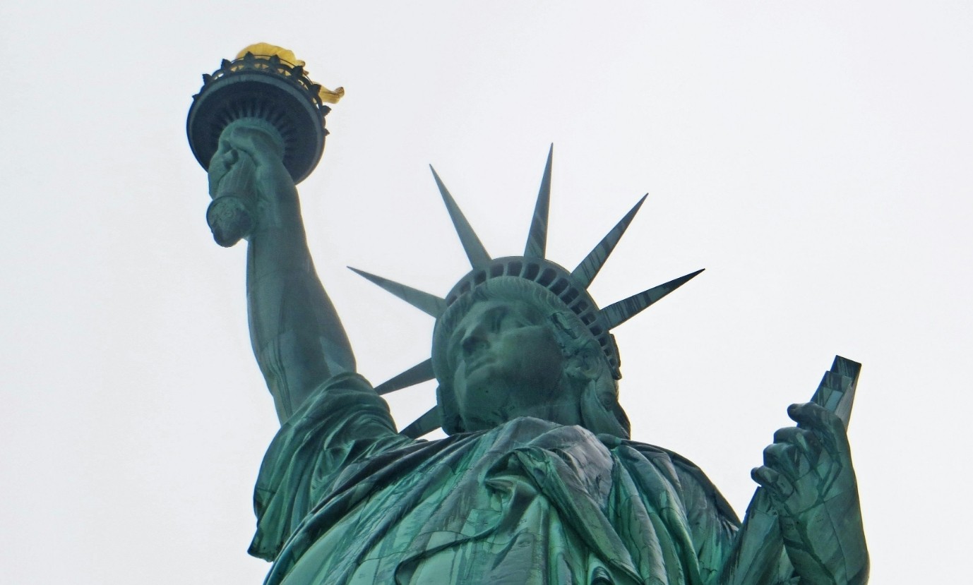 Estátua da Liberdade, em tom esverdeado, vista de baixo para cima, com a tocha dourada erguida na mão direita e a coroa de sete pontas na cabeça, contra um céu claro