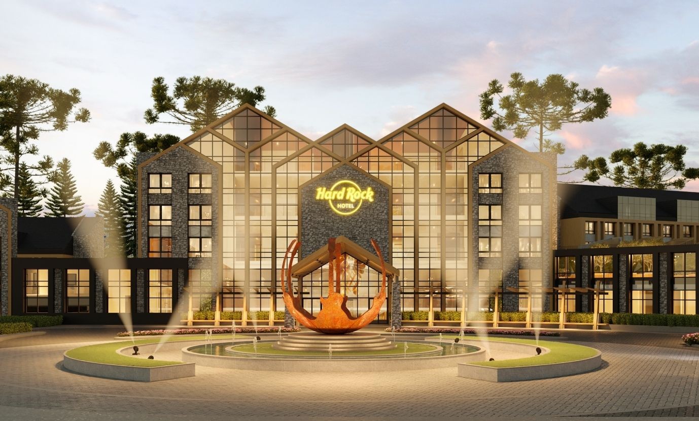 Fachada noturna do Hard Rock Hotel, com arquitetura moderna em vidro e pedra, cercado por araucárias. Em frente, uma fonte circular com uma escultura de guitarra estilizada e jatos de luz iluminando a entrada