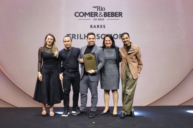 Cinco pessoas sorrindo no palco, o homem central de terno cinza segura um pr&ecirc;mio dourado. Ao fundo, um tel&atilde;o exibe 