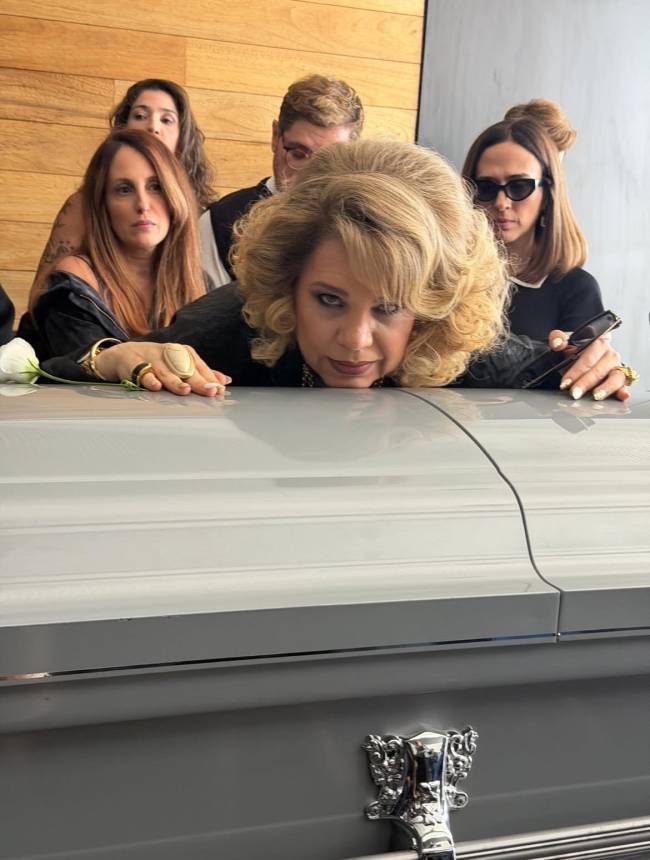 Mulher loira com cabelo volumoso e maquiagem forte, deitada sobre um caix&atilde;o cinza, com express&atilde;o s&eacute;ria. Atr&aacute;s dela, outras quatro pessoas, incluindo uma mulher de &oacute;culos escuros e um homem de terno, observam com semblantes tristes. Uma rosa branca est&aacute; no caix&atilde;o. O fundo &eacute; uma parede de madeira.