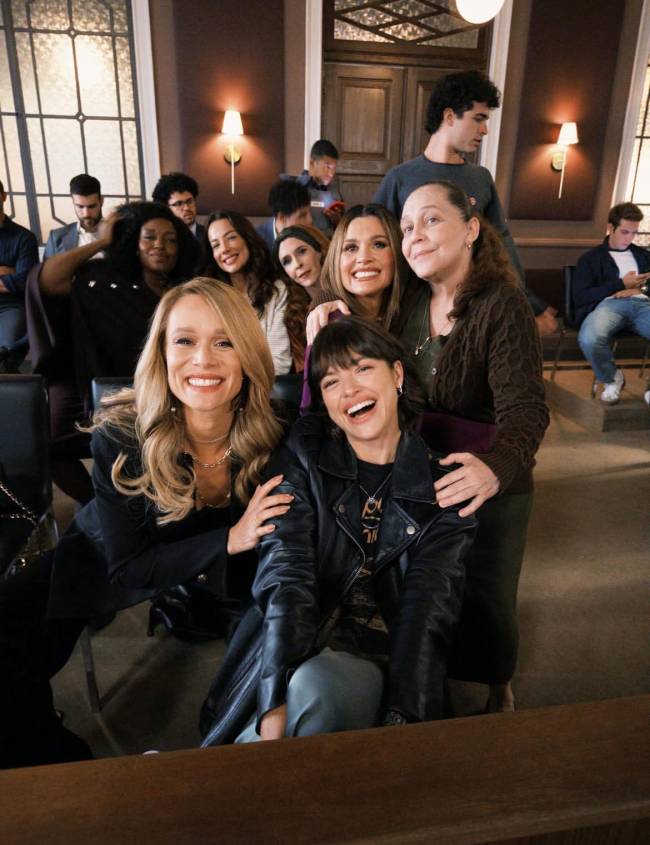 Grupo de mulheres sorrindo para a c&acirc;mera, com outras pessoas ao fundo em um ambiente que parece ser um tribunal ou sala de aula. A mulher loira na frente veste blazer preto, e a morena ao lado usa jaqueta de couro preta.