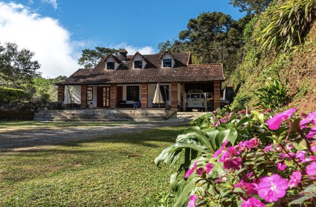 Casa de campo r&uacute;stica com telhado de cer&acirc;mica e paredes de madeira, cercada por gramado e &aacute;rvores. Um carro branco est&aacute; na garagem e flores roxas em primeiro plano.