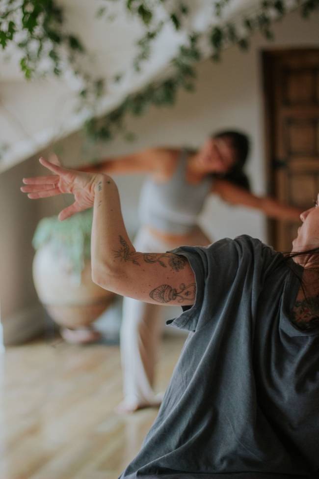 Duas mulheres praticando yoga ou dan&ccedil;a em um ambiente claro. A mulher em primeiro plano, de camiseta cinza e bra&ccedil;os tatuados, estende a m&atilde;o esquerda. Ao fundo, outra mulher de top cinza e cal&ccedil;a clara, com os bra&ccedil;os abertos, est&aacute; em movimento. Plantas penduradas e um vaso decoram o espa&ccedil;o