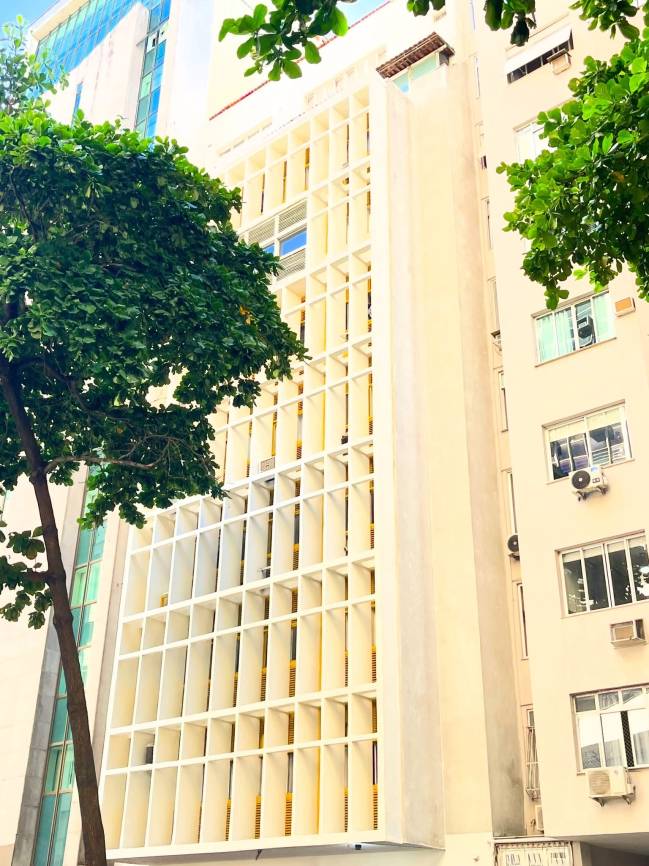 Fachada de pr&eacute;dio alto com brises verticais brancos e janelas amarelas, ladeado por outro edif&iacute;cio com janelas e aparelhos de ar condicionado, e &aacute;rvores com folhagem verde em primeiro plano
