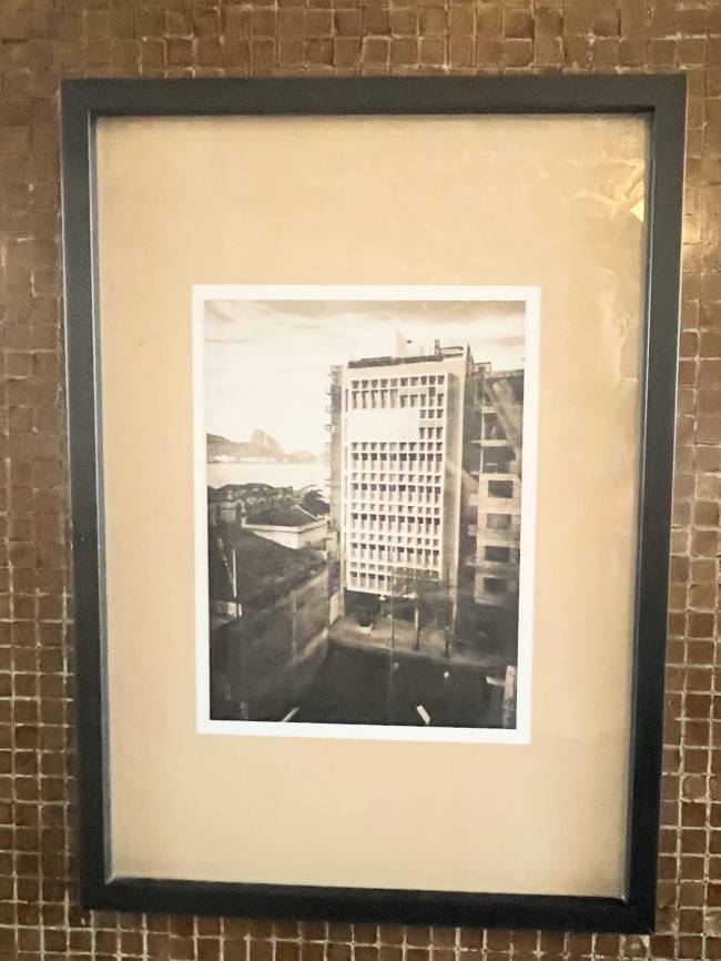 Fotografia em preto e branco de um pr&eacute;dio alto com muitas janelas, emoldurada em preto com passe-partout bege. Ao fundo, montanhas e o mar do Rio de Janeiro. A imagem est&aacute; pendurada em uma parede de azulejos marrons.