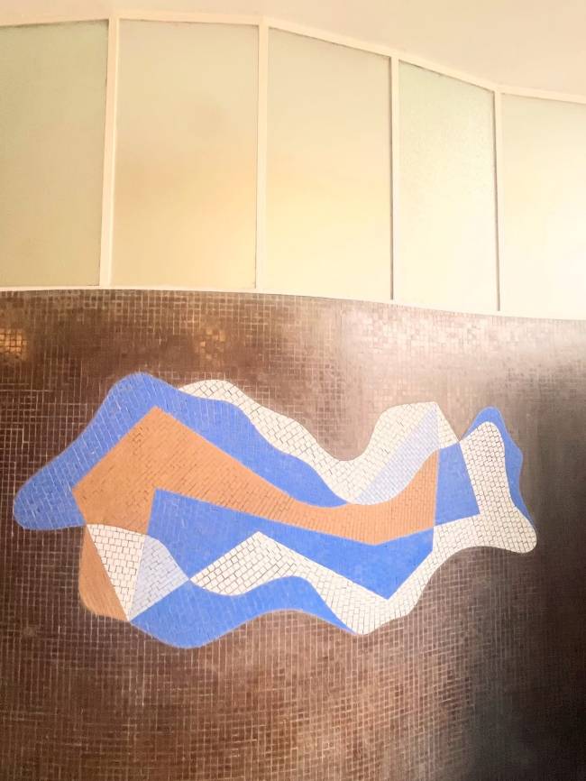 Mosaico abstrato em tons de azul, branco e laranja, com formas onduladas, sobre uma parede de pequenos azulejos marrons. Acima, janelas retangulares com vidro fosco e molduras brancas