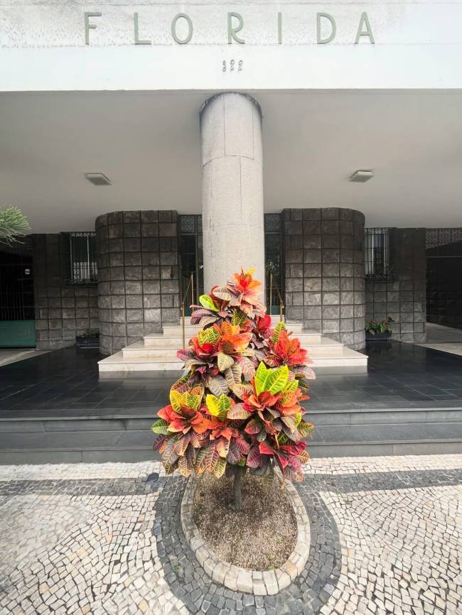 Entrada de um edif&iacute;cio com a palavra FLORIDA e o n&uacute;mero 122 em relevo na fachada. Um grande vaso de croton com folhas coloridas em tons de vermelho, laranja e verde est&aacute; em frente &agrave; porta, sobre um piso de pedras portuguesas