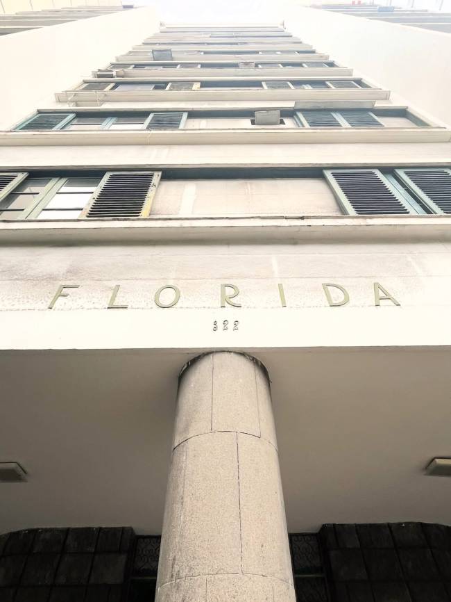 Vista de baixo para cima de um edif&iacute;cio branco com janelas e persianas verdes, algumas abertas. Na fachada, letras douradas formam FLORIDA e abaixo, o n&uacute;mero 322. Uma coluna cinza de concreto sustenta a estrutura, com ilumina&ccedil;&atilde;o no teto.